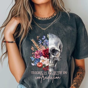 Puede incluir: Una camiseta negra con un diseño de calavera y flores. La calavera está dividida en dos, con flores a un lado y la calavera al otro. El texto "There is beauty in contradiction" está impreso debajo del diseño.