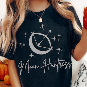 May include: Dark gray t-shirt featuring a crescent moon and arrow design with surrounding stars. The words "Moon Huntress" are written in a cursive font below the graphic. The shirt is a casual style.