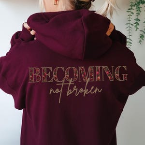 Może przedstawiać: Bordowa bluza z kapturem z napisem "BECOMING not broken" z tyłu. Słowo "BECOMING" jest w kwiatowym wzorze. Bluza z kapturem ma kaptur i sznurek. Tło jest jasne.