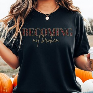 Może przedstawiać: Czarny t-shirt z napisem "BECOMING not broken". Słowo "BECOMING" ma kwiatowy wzór. Tekst jest w kroju szeryfowym. T-shirt ma krótkie rękawy.