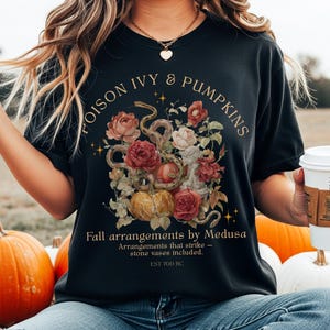 May include: Black t-shirt with a floral and snake design. The design includes pumpkins, roses, and the text "POISON IVY & PUMPKINS". Below the design is the text "Fall arrangements by Medusa" and "EST 700 BC".