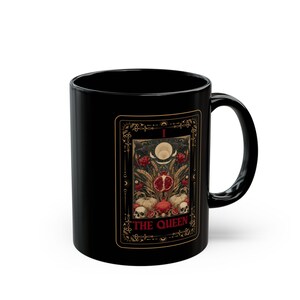 Puede incluir: Taza de cer&aacute;mica negra con dise&ntilde;o de carta de tarot. La carta representa una granada, calaveras y elementos bot&aacute;nicos. Las palabras "THE QUEEN" est&aacute;n impresas en la carta. La taza tiene un asa curva.