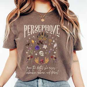 Puede incluir: Una camiseta marrón con un diseño gráfico que presenta la palabra "Persephone" en una fuente vintage, una corona y varios símbolos florales y otros. El texto debajo del gráfico dice "from the depths she rises - unbroken, reborn, and eternal."