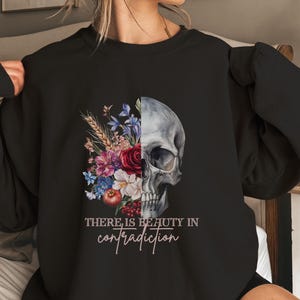 Puede incluir: Sudadera negra con un gráfico de una calavera con flores en un lado y el texto "THERE IS BEAUTY IN CONTRADICTION" en el otro.