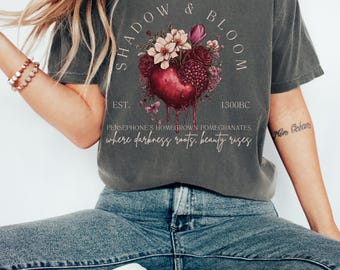 May include: A dark gray t-shirt with a graphic design of a red heart with flowers and the text "SHADOW & BLOOM EST. 1300BC PERSEPHONE'S HOMEGROWN POMEGRANATES where darkness roots, beauty rises"