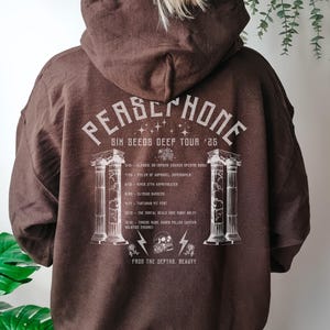 Puede incluir: Sudadera con capucha marrón con un estampado gráfico blanco de una banda llamada Persephone. El gráfico incluye el texto "Six Seeds Deef Tour '25" y una lista de fechas y lugares de la gira.
