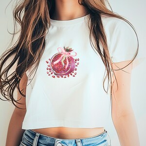 Puede incluir: Una camiseta blanca recortada con una cinta rosa y un gráfico de granada roja.