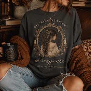 Puede incluir: Camiseta gris oscuro con una ilustración vintage de una mujer con cabello de serpiente, rodeada por un marco floral. El texto dice "SHE LEARNED TO SPEAK in serpents." También se ve una taza negra con "ROMANCE READER CLUB".
