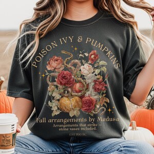 May include: Dark gray t-shirt with a floral and snake graphic. The design includes roses, pumpkins, and the text "POISON IVY & PUMPKINS" and "Fall arrangements by Medusa". The shirt also has the text "Arrangements that strike - stone vases included. EST 700 BC."