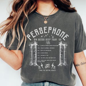 Puede incluir: Una camiseta gris oscuro con un diseño gráfico blanco que presenta el texto "PERSEPHONE SIX SEEDS DEEP TOUR '25" y una imagen estilizada de dos columnas con una calavera y rosas. Las fechas de la gira se enumeran en la camiseta.