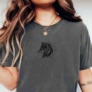 Puede incluir: Camiseta gris oscuro con un diseño de cabeza de lobo negro y el texto "echoes of the wild". La camiseta tiene cuello redondo y mangas cortas. La tela es suave y cómoda.