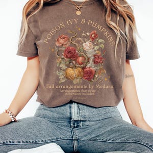 May include: Brown t-shirt with the text "POISON IVY & PUMPKINS" in an arch. Below the text is a floral arrangement with roses, pumpkins, and a snake. The text "Fall arrangements by Medusa" is below the floral arrangement.