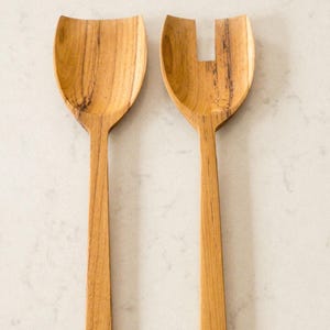 Puede incluir: Dos utensilios para ensalada de madera. Uno tiene forma de cuchara y el otro de tenedor. Ambos están hechos de madera de color marrón claro con vetas visibles. Los mangos son largos y delgados, y las cabezas para servir son anchas y curvadas.