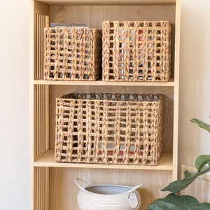 Può includere: Un'unità a scaffale in legno con quattro ripiani, ognuno dei quali contiene cesti intrecciati. Lo scaffale superiore ha un oggetto in ceramica bianca e una ciotola di legno. Lo scaffale inferiore ha un cesto intrecciato bianco con manici.