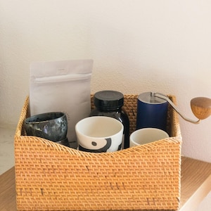 Può includere: Un cesto intrecciato in vimini con una tazza in ceramica marrone e nera, una tazza in ceramica bianca con pois neri, una tazza in ceramica bianca, un barattolo nero con un coperchio argentato, un macinacaffè blu con manico argentato e una busta bianca con una cerniera.