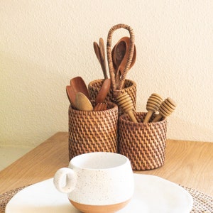 Könnte beinhalten: Eine weiße Keramiktasse mit braunen Flecken steht auf einem weißen Teller auf einem geflochtenen Untersetzer. Im Hintergrund stehen drei geflochtene Körbe gefüllt mit Holzlöffeln und einem Holzlöffel für Honig.