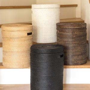 May include: Four cylindrical woven storage baskets in natural, white, dark brown, and black. Each has a lid and a handle cutout. The baskets are displayed on wooden stairs. Measurements are in cm.