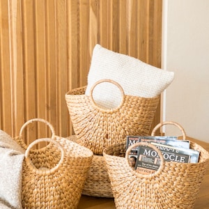 Peut inclure: Trois paniers en osier tressé avec des anses. Les paniers sont de différentes tailles et sont posés sur un sol en bois. Le plus grand panier contient un coussin blanc.