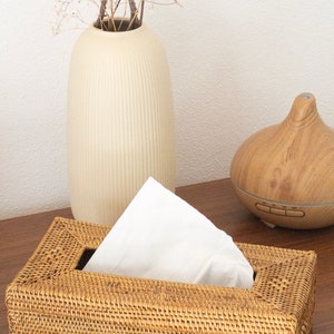 Pode incluir: Uma caixa de lenços retangular feita de rattan tecido, com uma pilha de lenços brancos visíveis. Um vaso creme com flores secas e um difusor de óleo essencial de madeira estão no fundo. A caixa de lenços está em uma superfície de madeira.