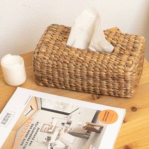 Könnte beinhalten: Ein geflochtener Weiden-Tischtuchhalter mit einem weißen Taschentuchspender im Inneren. Der Halter steht auf einem Holztisch mit einem Magazin darauf.