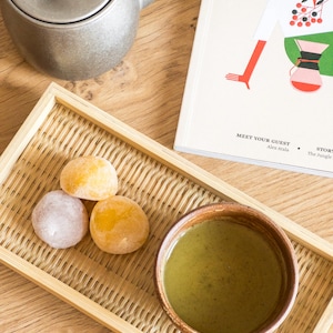 Può includere: Un vassoio di legno con un motivo in bambù intrecciato contiene una piccola ciotola di tè verde, tre mochi e una teiera d'argento. Il vassoio è su un tavolo di legno accanto a un libro intitolato "Meet Your Guest" di Alex Atala.