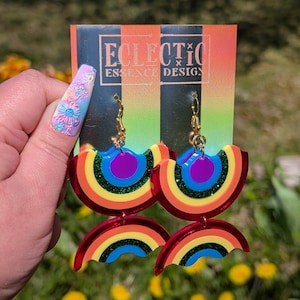 May include: A pair of dangle earrings featuring two stacked rainbow-shaped acrylic pieces in red, orange, yellow, green, blue, and purple with glitter accents. The earrings are attached to a display card labelled ECLECTIC ESSENCE DESIGN.