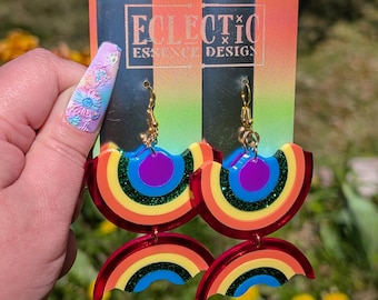 Puede incluir: Pendientes colgantes con dos piezas de acrílico en forma de arcoíris superpuestas en rojo, naranja, amarillo, verde, azul y morado. El diseño incluye una banda verde con purpurina y ganchos de metal dorado. Presentados en una tarjeta de marca.