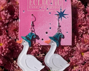 Puede incluir: Un par de pendientes colgantes con forma de gansos blancos con sombreros de bruja azul verdoso y picos naranjas, sobre bases verdes. Los pendientes están sujetos a una tarjeta rosa con el texto ECLECTIC ESSENCE DESIGN y una estrella.