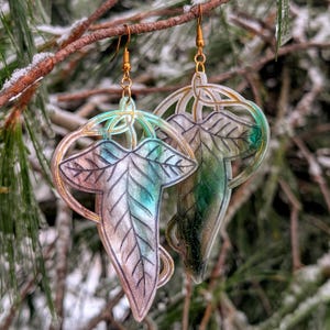 May include: A pair of leaf-shaped earrings with gold-colored hooks. The earrings have a translucent design with shades of green, blue, and pink. They are hanging from a tree branch with a snowy background.