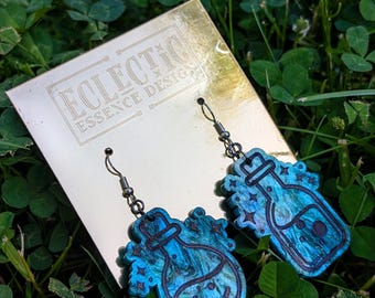 Peut inclure: Une paire de boucles d'oreilles pendantes en forme de flacons de potion bleu et turquoise avec des détails gravés d'étoiles et de bulles. Elles sont fixées sur un carton de présentation doré portant le texte ECLECTIC ESSENCE DESIGN en noir.