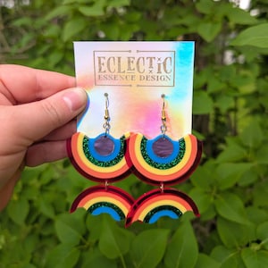 May include: Rainbow-themed earrings with gold-colored hooks. The earrings feature a layered design with red, orange, yellow, green, and blue arcs. The earrings are displayed on a holographic card with the text "Eclectic Essence Design".