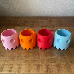 Puede incluir: Cuatro pequeños maceteros con forma de fantasma en rosa, naranja, rojo y azul.