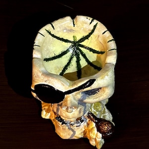 Puede incluir: Un cenicero con forma de calavera con un parche negro en el ojo y una pipa. El interior del cenicero es amarillo con un diseño de telaraña verde. La calavera es blanquecina con sombras oscuras, lo que le da un aspecto envejecido.
