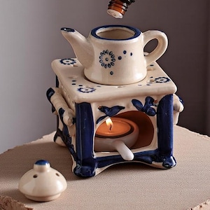 Op de afbeelding: Een keramische etherische olieverspreider met een theepotontwerp. De wit en blauw geschilderde diffuser heeft een kleine theepot bovenop en een kaarsje binnenin. Een hand giet olie uit een kleine bruine fles in de theepot.