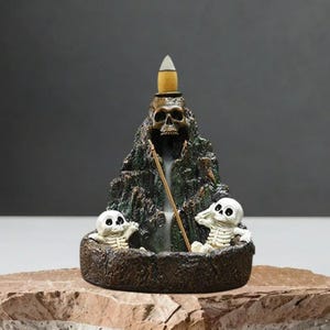Puede incluir: Un quemador de incienso decorativo con forma de montaña con calavera, con dos pequeños esqueletos en la base. El quemador es marrón y verde, con un diseño detallado y texturizado. Un cono de incienso arde en la parte superior.