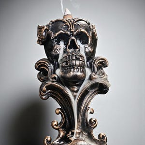 Puede incluir: Un quemador de incienso decorativo con forma de calavera en negro y bronce. La calavera tiene detalles intrincados y emite humo. La base presenta diseños ornamentados y arremolinados. Un cono de incienso está en la parte superior.