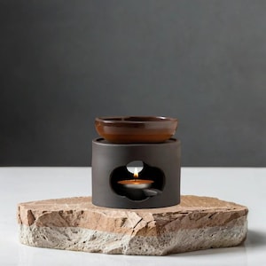 Puede incluir: Un difusor de aceites esenciales de cerámica gris oscuro con un cuenco marrón. Una pequeña vela arde en el interior, emitiendo un brillo cálido. El difusor se encuentra sobre una base de piedra texturizada y multicolor.