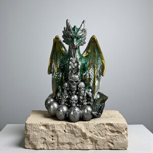 Puede incluir: Figura detallada de un dragón con escamas verdes y plateadas, alas con bordes dorados y una colección de calaveras. El dragón está posado sobre un grupo de huevos plateados agrietados. La figura se exhibe sobre un bloque de piedra clara texturizada.
