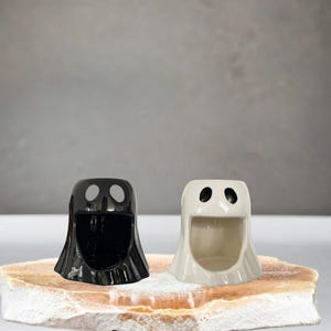 May include: Two ceramic ghost-shaped holders, one black and one off-white, each with two eye-shaped openings. The holders have a wide opening at the bottom. They are displayed on a light brown, textured surface.