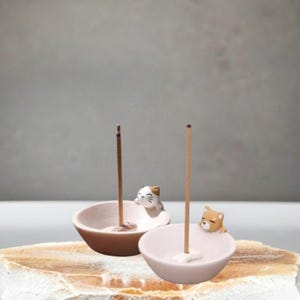Op de afbeelding: Twee keramische wierookhouders, elk met een klein dierenbeeldje. Eén heeft een kat, de andere een hond. Wierookstokjes worden in de houders gestoken. De houders zijn lichtroze en bruin.