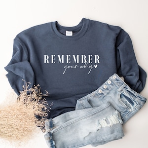 May include: A navy blue sweatshirt with the text "REMEMBER your why" in white. The sweatshirt is paired with light blue denim jeans. The image is taken on a white background.