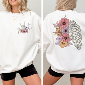 Op de afbeelding: Witte trui met een bloemenontwerp en de tekst "p i v o t" op de voorkant. De achterkant van de trui heeft een bloemenontwerp met een ribbenkast.