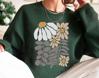 May include: A dark forest green crewneck sweatshirt featuring a central graphic print of stylized flowers and leaves in shades of white, beige, mustard yellow, and muted mauve. The long-sleeved garment has a ribbed neckline and cuffs.