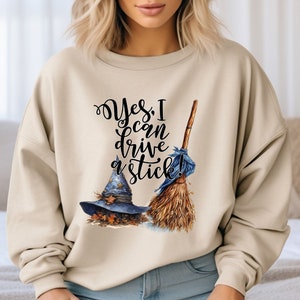 May include: A beige crewneck sweatshirt featuring a graphic print with the text "Yes, I can drive a stick!" in black script. The design includes a blue pointed witch hat adorned with gold stars and autumn leaves, alongside a wooden broomstick.