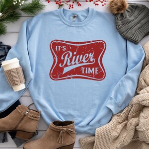 May include: A light blue crewneck sweatshirt featuring a red, distressed-style rectangular graphic with rounded corners. The graphic contains the light blue text "IT'S River TIME" in a stylized font. The sweatshirt has long sleeves and a ribbed collar.