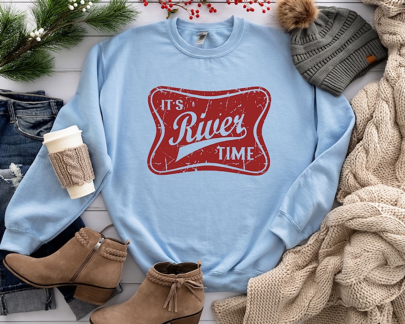 May include: A light blue crewneck sweatshirt featuring a red, distressed-style rectangular graphic with rounded corners. The graphic contains the light blue text "IT'S River TIME" in a stylized font. The sweatshirt has long sleeves and a ribbed collar.