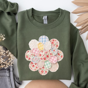 May include: An olive green sweatshirt featuring a large, appliqued flower design. The flower is composed of various fabric patches in floral, gingham, and solid patterns. The sweatshirt has long sleeves and a crew neck.