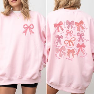 May include: A light pink crewneck sweatshirt featuring a small pink bow graphic on the front left chest. The back displays a large collection of various pink bow illustrations accompanied by the words Amazing, Valued, Brave, Powerful, Important, Strong, Unique, Loved, Precious, and Protected.