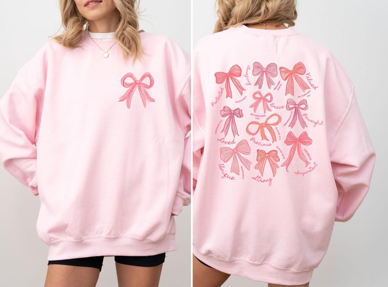 May include: A light pink crewneck sweatshirt featuring a small pink bow graphic on the front left chest. The back displays a large collection of various pink bow illustrations accompanied by the words Amazing, Valued, Brave, Powerful, Important, Strong, Unique, Loved, Precious, and Protected.