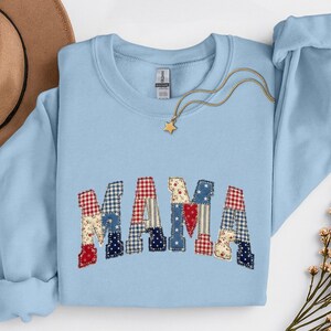 May include: Light blue sweatshirt with the word "MAMA" appliqued across the front. The letters are made of patchwork fabric in red, white, and blue patterns. A gold necklace with a star pendant is draped on the sweatshirt.